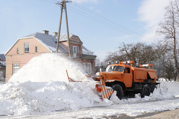 A large car with a plow clears the road from snow. Orange cargo special equipment is struggling with the elements in winter. Removing the effects of the snowstorm. Difficulties in traffic.Frozen water
