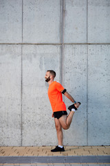 Fototapeta na wymiar Sporty man in black and orange sportswear stretching leg and standing next to wall on the street.