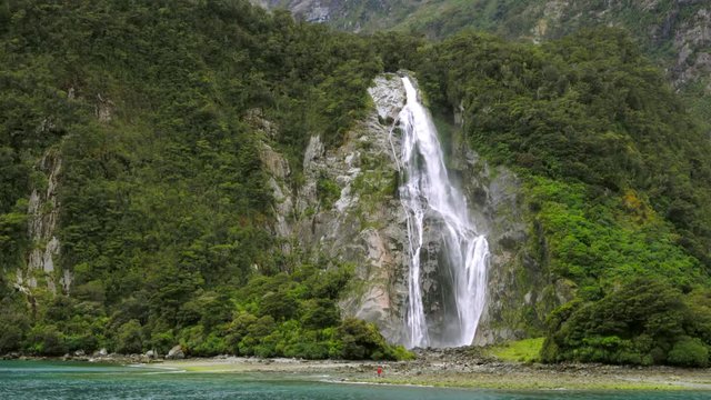 Handheld shot from boat sailing past waterfall on side of mountain slope overgrown with lush greenery