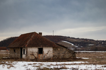 Old small abandoned house on meadow.Winter rural scene at sunset with small bits of snow.