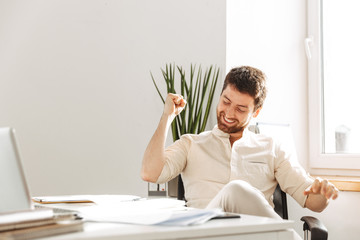 Photo of handsome businessman 30s wearing white shirt rejoicing, while working in bright office