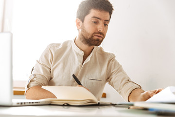 Image of handsome businessman 30s wearing white shirt working with laptop and paper documents, while sitting in bright office
