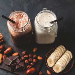 Banana and chocolate smoothie in two jars, protein milkshake, nuts, chocolate cubes, banana slices, dates, cinnamon sticks, healthy eating on a dark background