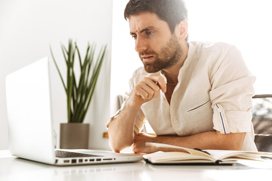 Image of successful businesslike man 30s wearing white shirt working with laptop and paper documents, while sitting in bright office