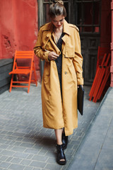 Young beautiful smiling woman in trench coat and boots holding bag in hand while spending time in old cozy courtyard