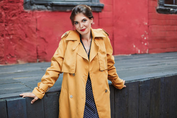 Young beautiful smiling woman in orange trench coat and black polka dot dress dreamily looking in camera while spending time alone in old courtyard