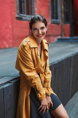 Young beautiful joyful woman in orange trench coat and black polka dot dress happily looking in camera while spending time alone in old courtyard