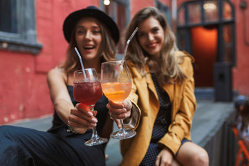 Young beautiful cheerful girls holding cocktails in hands happily looking in camera while spending time together in old cozy courtyard of cafe