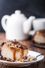 Close up view of a piece of peanut butter chocolate caramel cheesecake pie and cup of coffee on wooden background with a coffee pot and cheesecake in the background. A Sweet Coffee Break concept 