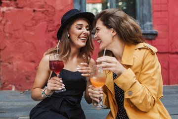 Young beautiful laughing girls holding cocktails in hands happily looking at each other while spending time together in old cozy courtyard of cafe