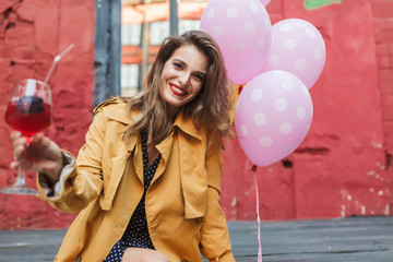 Young beautiful smiling woman in orange trench coat holding cocktail in hand happily looking in...