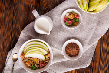 Breakfast bowl of oat granola with apple and dry fruit in a white bowl on brown wooden background  with a yogurt. Copy space