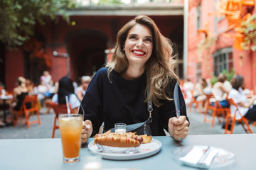 Young cheerful woman in black dress holding fork and knife in hands with food on table joyfully looking aside while spending time in cozy courtyard of cafe