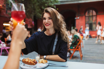 Young beautiful smiling woman in black dress sitting at the table with food holding cocktail in hand while happily looking at friend in old cozy courtyard of cafe