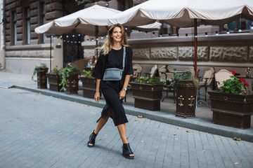 Young gorgeous smiling woman in black dress with cross bag joyfully looking in camera while walking on beautiful cozy city street alone