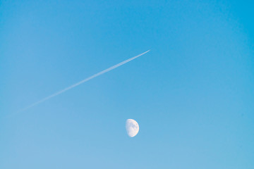 Condensation track of jet above moon in clear blue day sky. Minimalist blue background. Plane flies up diagonally. Airplane is flying in air space. White moon in cyan sky.