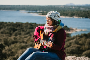 .Young woman sitting in the middle of the mountain overlooking a lake, playing her guitar. Playing music on a sunny and windy winter day. Lifestyle.
