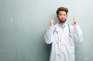 Young friendly doctor man against a grunge wall with a copy space crossing his fingers, wishes to be lucky for future projects, excited but worried, nervous expression closing eyes