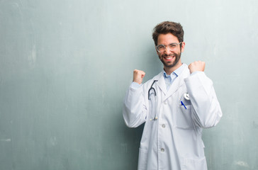 Young friendly doctor man against a grunge wall with a copy space very happy and excited, raising arms, celebrating a victory or success, winning the lottery