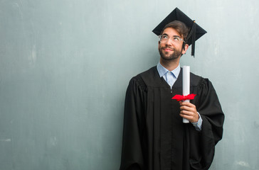 Young graduated man against a grunge wall with a copy space looking up, thinking of something fun and having an idea, concept of imagination, happy and excited