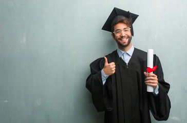 Young graduated man against a grunge wall with a copy space cheerful and excited, smiling and raising her thumb up, concept of success and approval, ok gesture