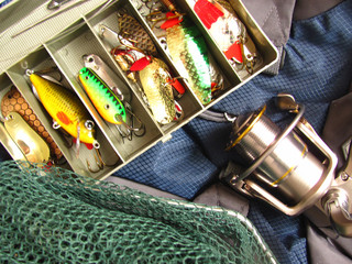 Set of colored spoon-baits, fishing net and spinning reel is on backpack background. Top view.