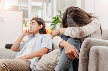 Fototapeta premium sadness young asian wife having quarrel and sitting on sofa after fight with husband behind her in house interior together, upset couple, love, divorce couple, family issues and relationship concept