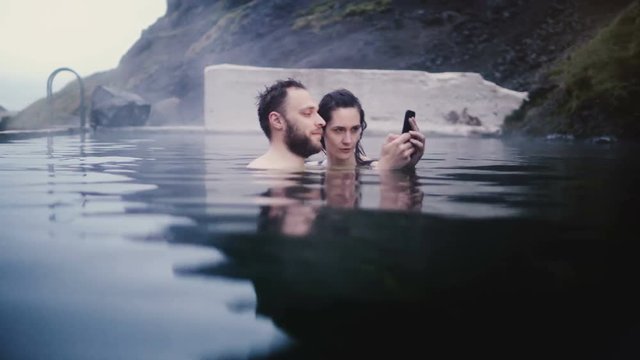 Young beautiful couple swimming in hot springs in Iceland. Traveling man and woman taking selfie photos on smartphone.