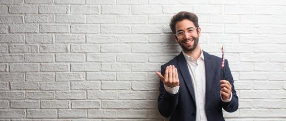 Young business man wearing a suit against a white bricks wall inviting to come, confident and smiling making a gesture with hand, being positive and friendly