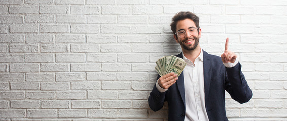 Young business man wearing a suit against a white bricks wall showing number one, symbol of...