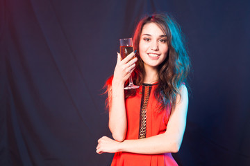 Naklejka premium Alcohol, people and party concept - Young celebrating woman in red dress. Beautiful model hold wine glass on dark background with copy space
