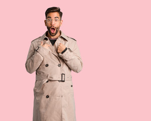 Young man wearing trench coat surprised, feels successful and prosperous