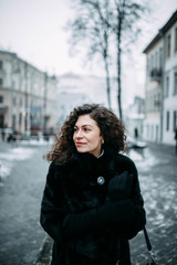 beautiful curly brunette girl in a fur black coat walks through the winter city.