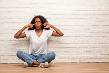 Young black woman sitting on a wooden floor covering ears with hands, angry and tired of hearing some sound