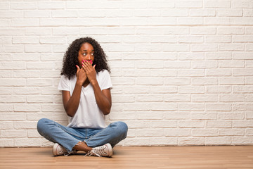 Young black woman sitting on a wooden floor covering mouth, symbol of silence and repression, trying not to say anything