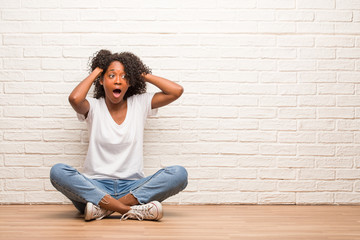 Young black woman sitting on a wooden floor worried and overwhelmed, forgetful, realize something,...