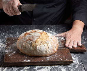 male chef hand holds knife over a whole round baked loaf of bread