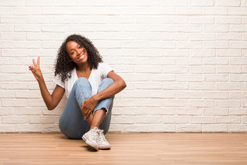 Young black woman sitting on wooden floor fun and happy, positive and natural, makes a gesture of victory, peace concept