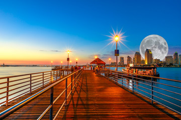 Scenic twiligtht view of Coronado wooden pier with docked ferry boat on Coronado Island, California, USA. San Diego skyline and urban downtown cityscape with big full moon over San Diego Bay. - obrazy, fototapety, plakaty