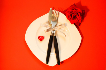 White asymmetrical plate, white red heart on it plus knife and fork with silver ribbon, red rose next to plate, red background. Perfect for Valentine's, anniversary, wedding, romantic event