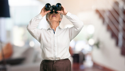 Beautiful senior woman surprised and amazed, looking with binoculars in the distance something interesting, concept of future opportunity at home.