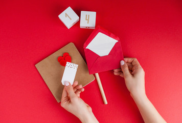 Female hands sign a card on a red background. Valentine's day concept. Valentine, hearts, pencil, wooden cubes with numbers.