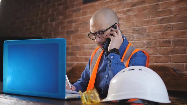 The architect on stroyploshadka works behind the laptop with the technical documentation and talking on the phone.