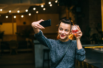 Party, technology and holidays concept - happy smiling young pretty woman drinking scotch whiskey glass taking selfie at night club or pub