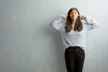 Young indian woman against a grunge wall covering ears with hands, angry and tired of hearing some...