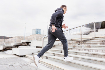 Young slim man a jogger in a gray tracksuit and sneakers moves up the steps around the sports stadium in early cloudy summer morning. Concept of energy and aerobic exercise. Copyspace