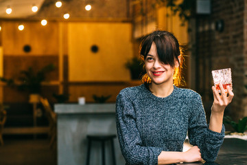 Drunk brunette student woman alone in happy smiling face expression holding and looking thoughtful to scotch whiskey glass at bar or pub in alcohol abuse and alcoholic concept.