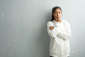 Young sporty indian woman against a gym wall thinking and looking up, confused about an idea, would be trying to find a solution