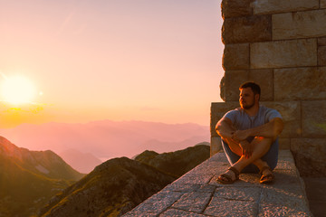 young adult man sitting on stone wall with beautiful view of sunset in mountains