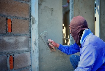 Plasterer wears balaclava using trowel to plastering concrete wall in construction site, side view with copy space